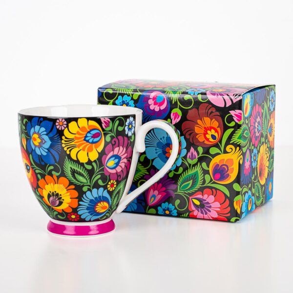 A vibrant Polish Folk Art Mug featuring hand-painted traditional Łowicz floral designs in bright colors on a black ceramic background, shown alongside a matching decorative box with the same colorful floral pattern. The mug has a pink base.