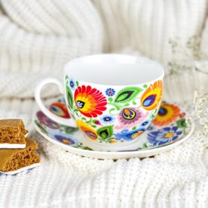 A vibrant ceramic teacup and saucer set adorned with colorful floral patterns inspired by Polish folk art. The teacup features a comfortable handle and is displayed alongside some gingerbread cookies on a cozy white knitted blanket.