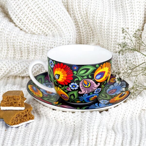A vibrant ceramic teacup and saucer set adorned with colorful floral patterns inspired by Polish folk art. The teacup features a comfortable handle and is displayed alongside some gingerbread cookies on a cozy white knitted blanket.